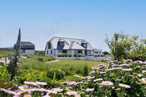 Une maison moderne à deux étages, dotée d’un toit en pente, entourée d’un jardin bien entretenu où poussent diverses plantes et fleurs.