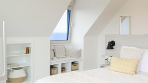 Une chambre avec un plafond en pente, une fenêtre avec des rideaux bleus, un lit avec une literie blanche, une table de chevet avec une lampe et des étagères de rangement avec des paniers.