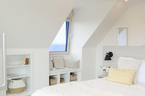 A bedroom with a sloped ceiling, a window with blue curtains, a bed with white bedding, a bedside table with a lamp, and storage shelves with baskets. - 5 Bed in Sennen (oc-b622152) (Trewolla)