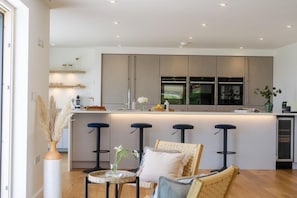 A modern kitchen with a central island, bar stools, and a dining area with a round table and chairs. - 5 Bed in Sennen (oc-b622152) (Trewolla)