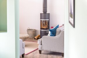 A cozy living room with a grey sofa, a wooden floor, a woven basket, and a wood-burning stove. - 3 Bed in Widemouth Bay (oc-b584375) (Widemouth Bay)