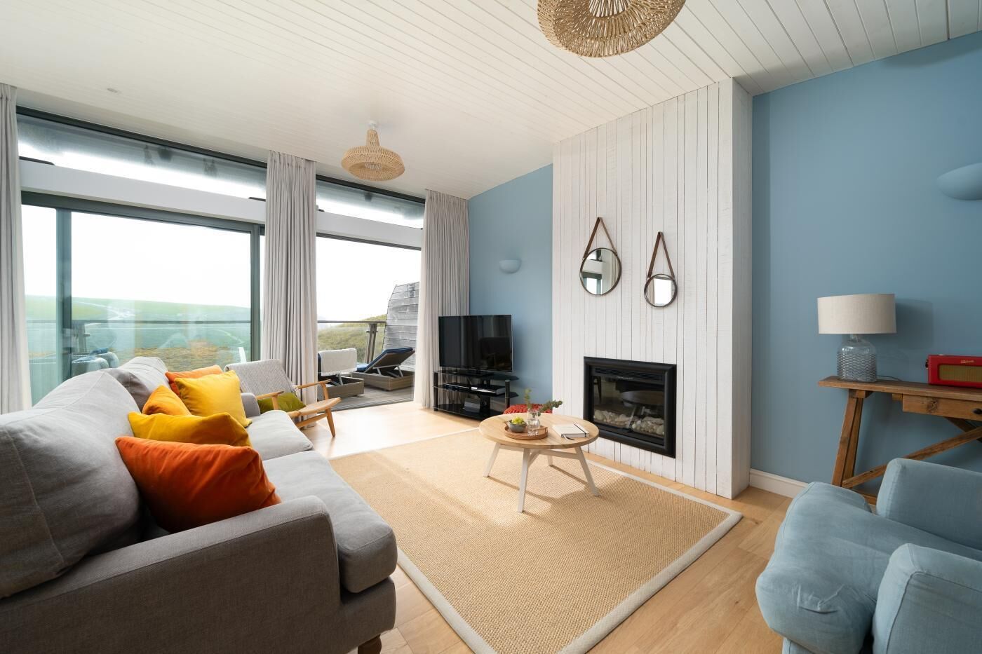 A modern living room with a sofa, armchairs, a TV, a fireplace, and a large window offering a view.