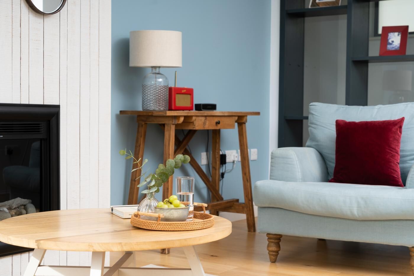 Un salon avec une table ronde en bois, un canapé bleu clair agrémenté d’un coussin rouge, un présentoir en bois avec une lampe et une cheminée.