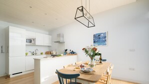 A modern kitchen with white cabinetry, a central island, and a dining area with a wooden table and chairs. - 2 Bed in Watergate Bay (oc-b584380) (Tregurrian)