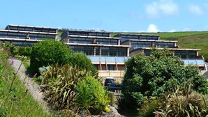 A modern multi-story building with a series of balconies and large windows, surrounded by lush greenery and a clear sky. - 2 Bed in Watergate Bay (oc-b584380) (Tregurrian)