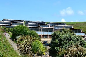 Ein modernes, mehrstöckiges Gebäude mit mehreren Balkonen und großen Fenstern, umgeben von üppigem Grün und blauem Himmel.