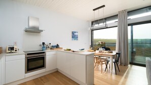 A modern kitchen with white cabinetry, a built-in oven, and a dining area with a table and chairs. - 2 Bed in Watergate Bay (oc-b584380) (Tregurrian)