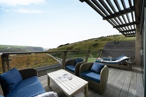A covered outdoor seating area with wicker furniture and a table, overlooking a hilly landscape. - 2 Bed in Watergate Bay (oc-b584380) (Tregurrian)