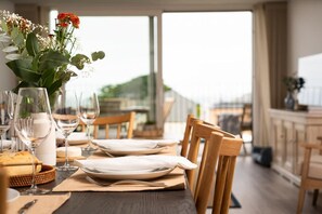 Ein Esstisch ist gedeckt mit Tellern, Besteck und Weingläsern, auf der linken Seite steht ein Blumenstrauß in einer Vase.