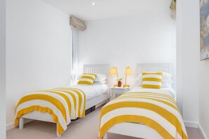 A hotel room with two beds, each with striped bedding, a nightstand with lamps, and a window with blinds. - 2 Bed in Porthtowan (oc-b584420) (Porthtowan)
