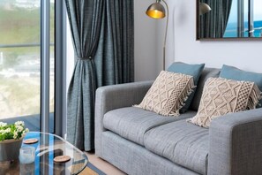 A modern living room with a grey sofa, macrame pillows, a floor lamp, and a view through large windows. - 2 Bed in Porthtowan (oc-b584420) (Porthtowan)