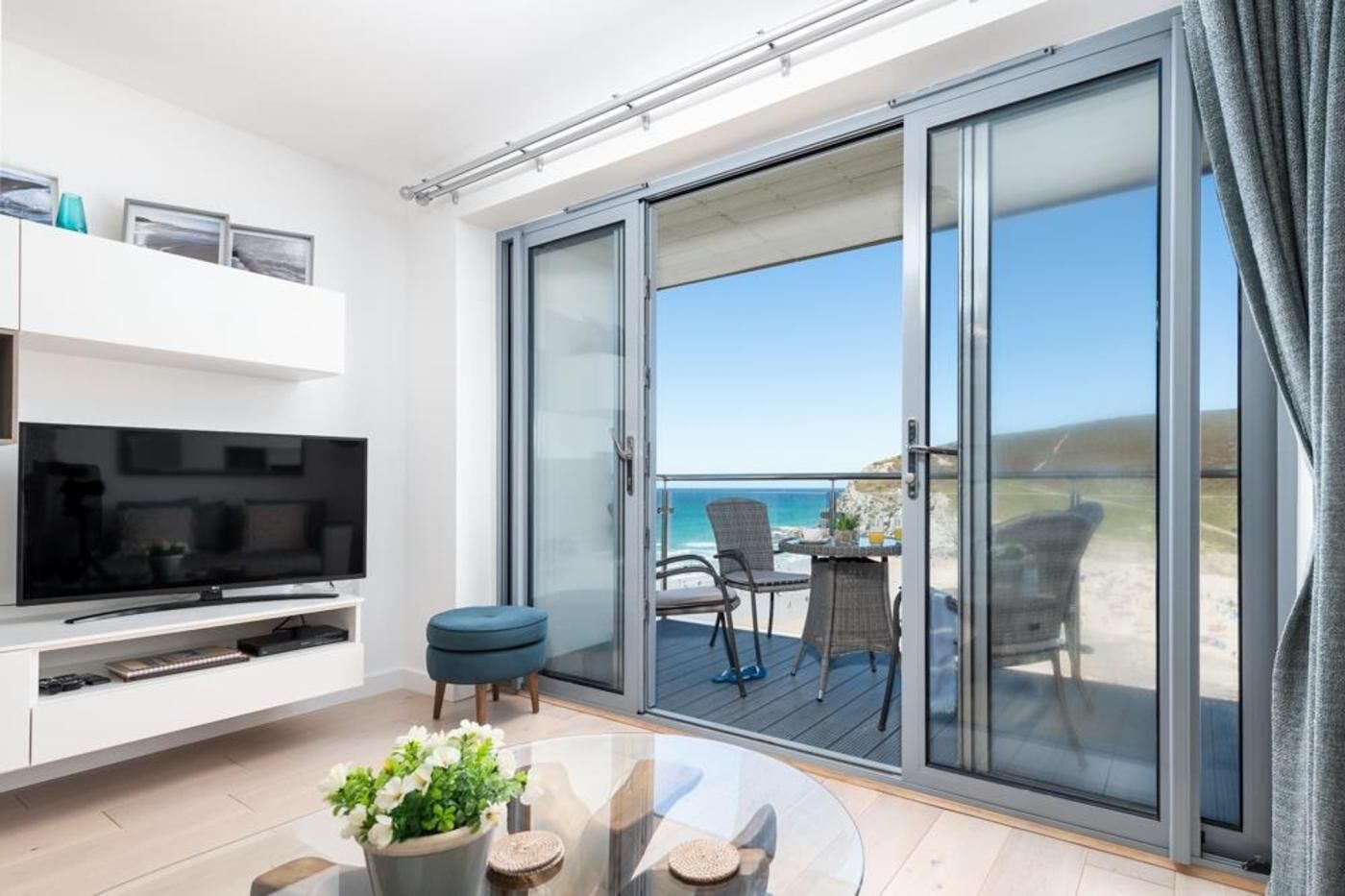 Ein modernes Wohnzimmer mit einem Flachbildfernseher, einem Glastisch mit Blumen und einer Schiebetür zu einem Balkon mit Blick auf den Strand.