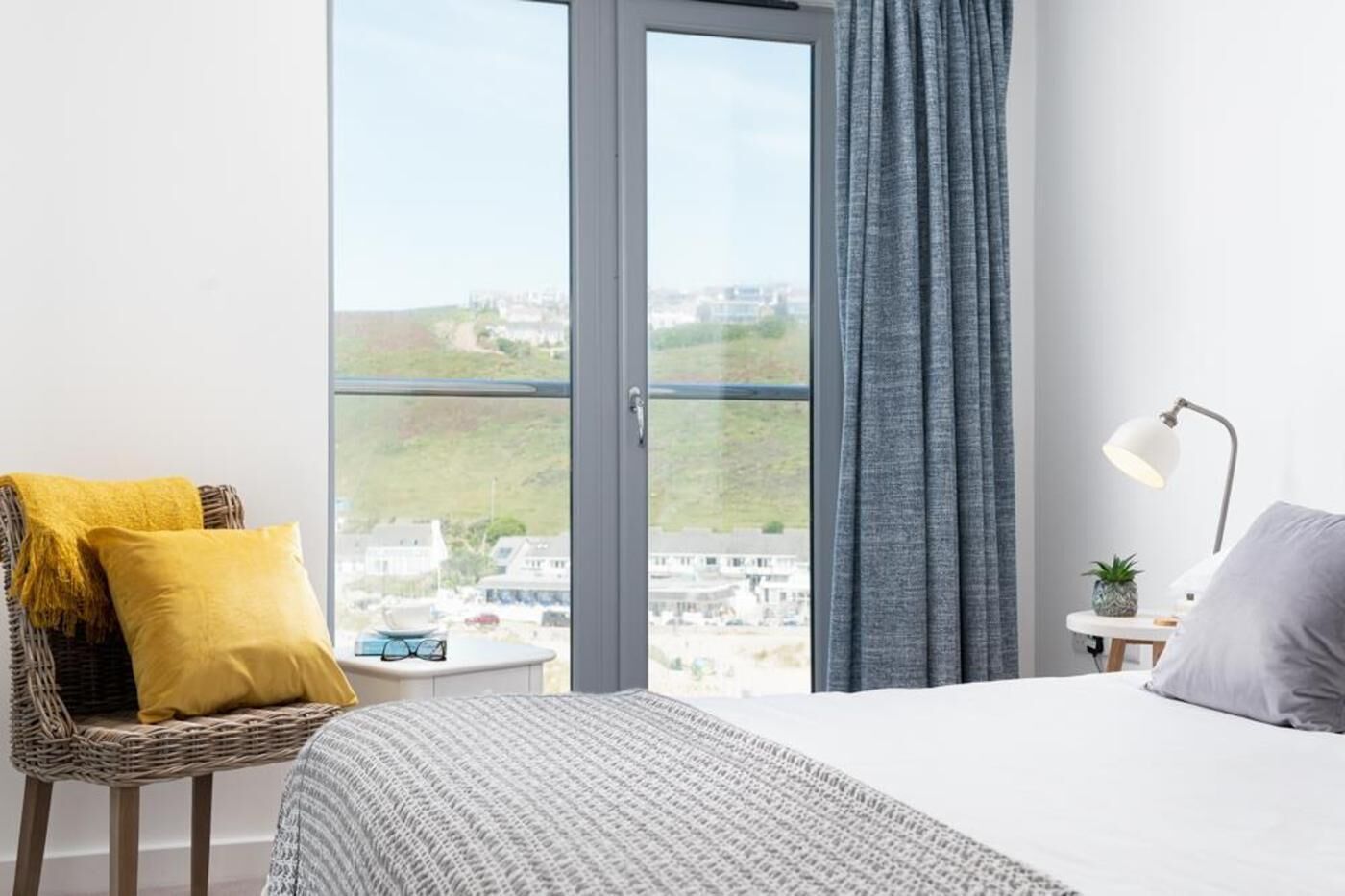 A hotel room with a bed, a chair with cushions, a nightstand with a lamp, and a view of a cityscape through a large window.