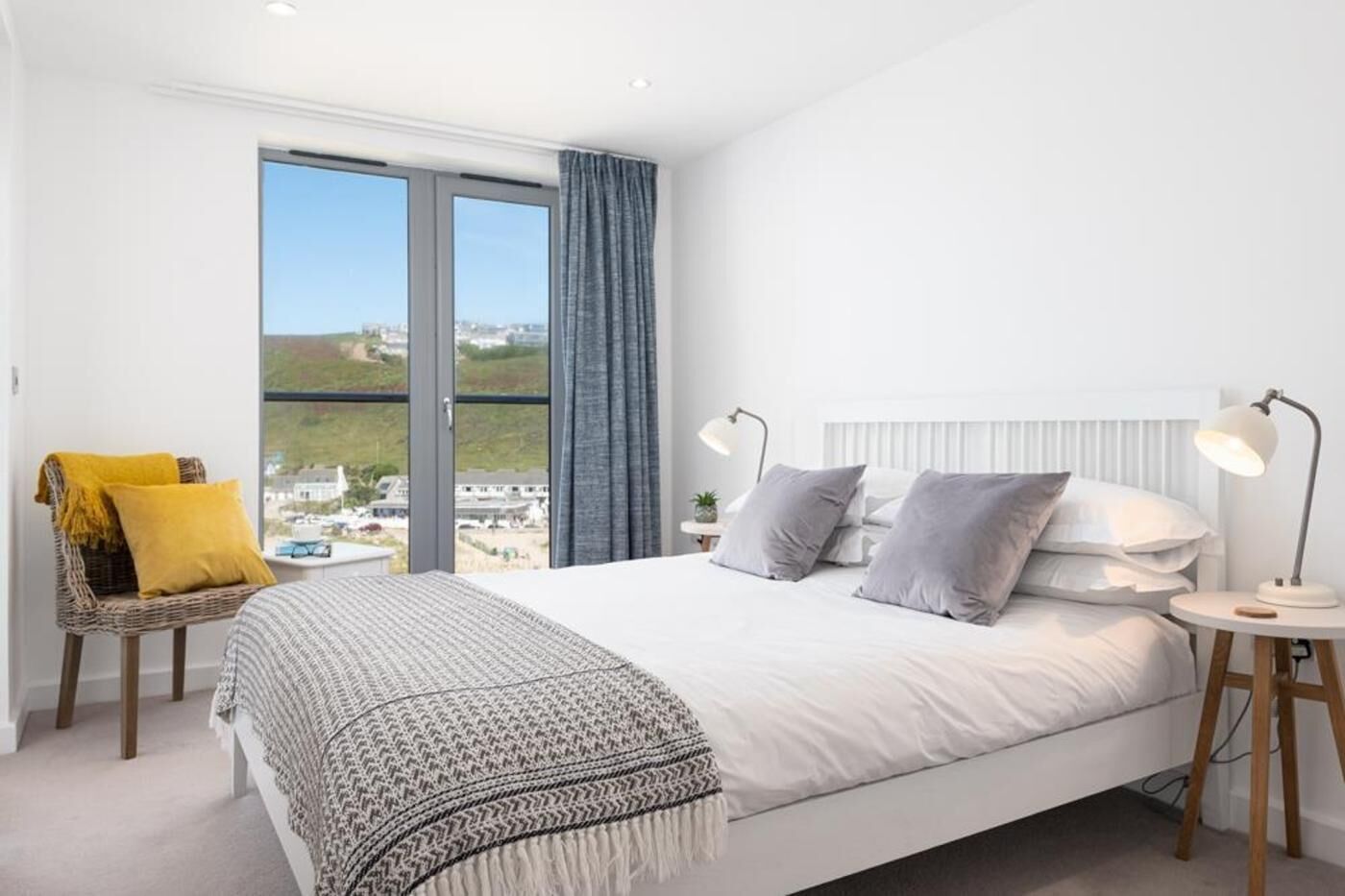 A bedroom with a large bed, two bedside lamps, a chair with a cushion, and a view of the city through a window.