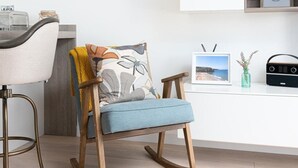 Una mecedora con cojín y almohada estampada, una radio y obras de arte enmarcadas sobre una pared blanca.