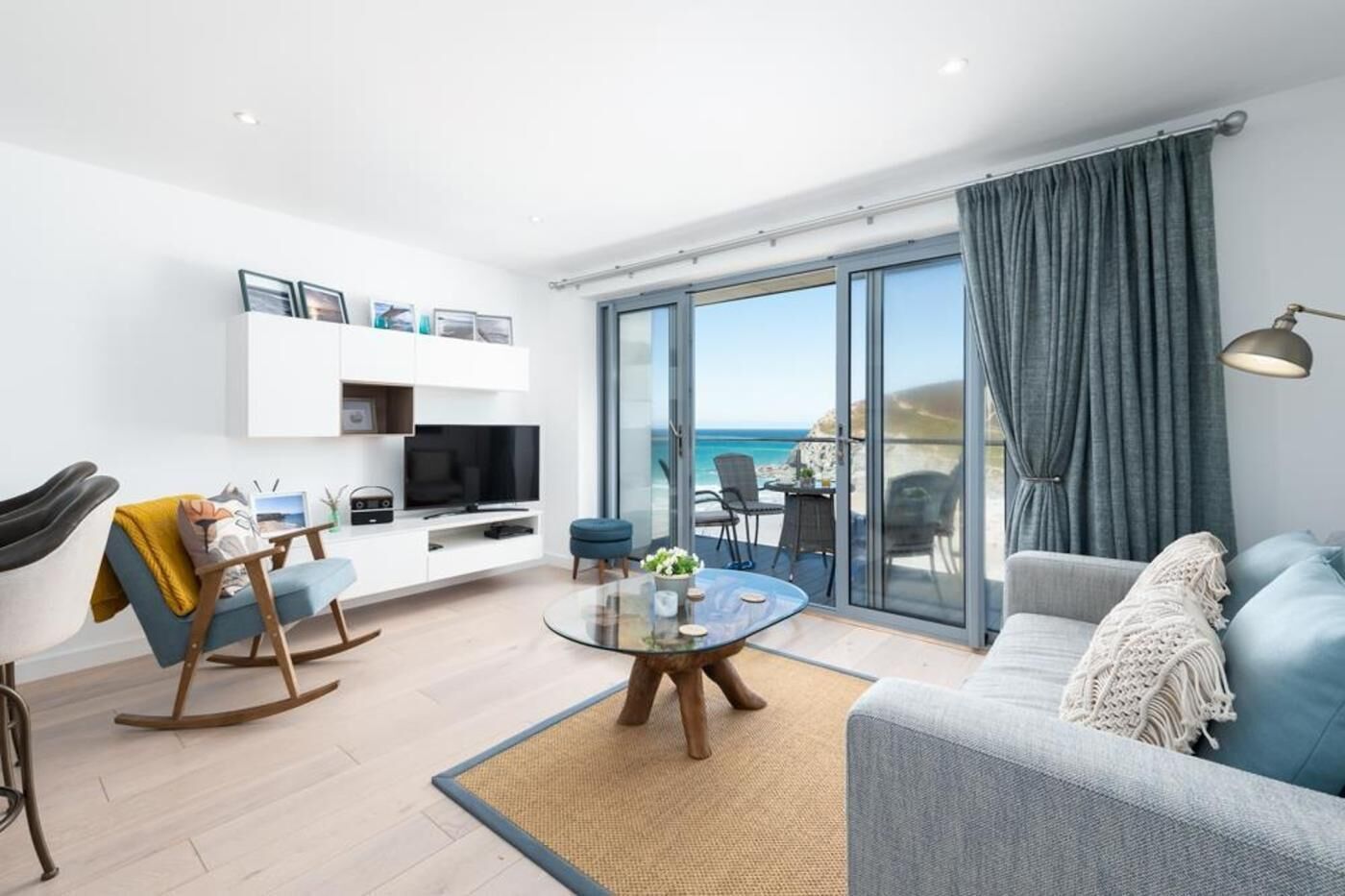 A modern living room with a sofa, a coffee table, and a TV. There is a balcony with a view of the sea.