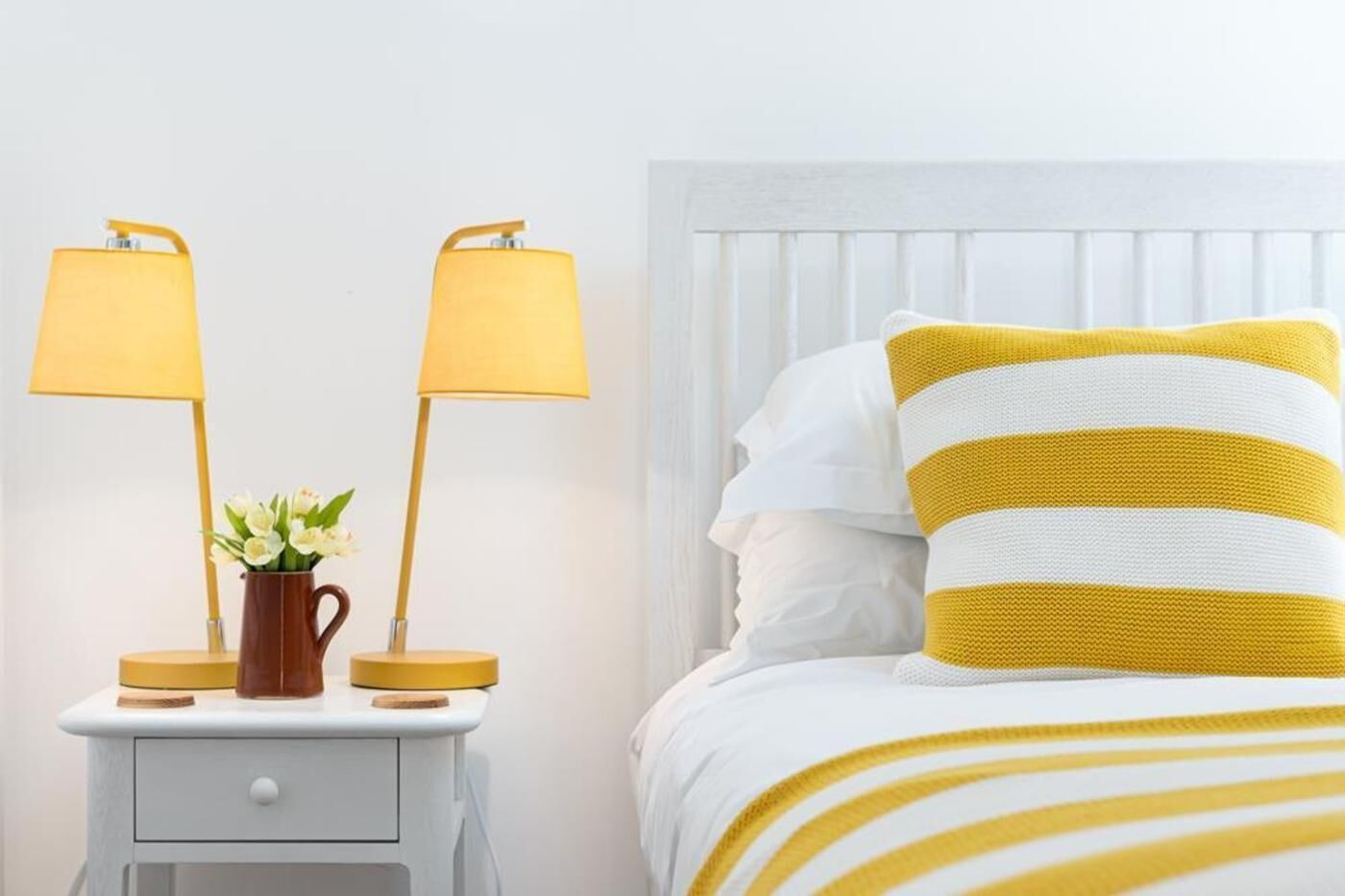 A bed with white and yellow striped bedding, a nightstand with two lamps, and a vase with flowers.
