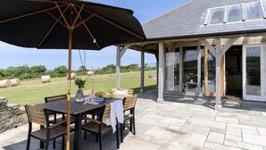An outdoor patio with a table and chairs, a large umbrella, and a view of a house with a covered porch and glass doors. - 3 Bed in Widemouth Bay (oc-b581851) (Rosecare)