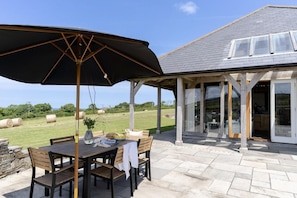 Eine Terrasse im Freien mit Tisch und Stühlen, einem großen Sonnenschirm und Blick auf ein Haus mit überdachter Veranda und Glasüren.