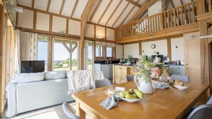 A spacious living area with a wooden ceiling, a dining table with a vase of flowers, and a view of the outdoors. - 3 Bed in Widemouth Bay (oc-b581851) (Rosecare)