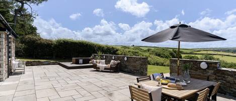 An outdoor patio with stone flooring, a wooden table with chairs, a sofa, and an umbrella.