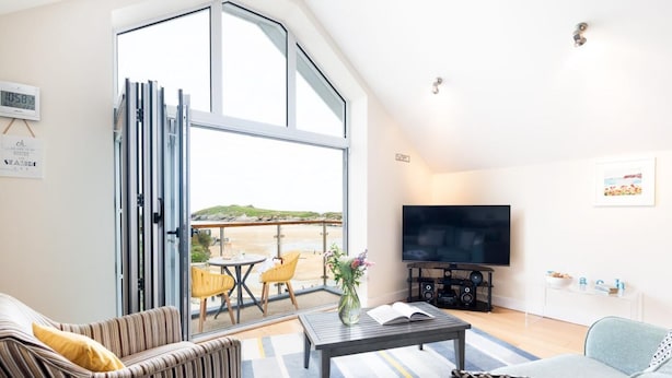 A modern living room with a large TV, seating area, and a view of the beach. - 2 Bed in Porth (oc-b584286) (Newquay)