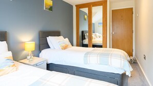 A bedroom with two beds, a bedside table with a lamp, a wardrobe, and a mirror. - 2 Bed in Porth (oc-b584286) (Newquay)