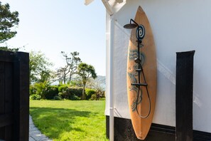 Une pomme de douche en forme de planche de surf en bois, fixée à un mur, avec vue sur une cour verdoyante et des arbres.