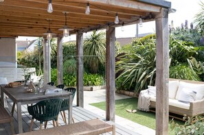 Een terras buiten met een houten tafel, stoelen en een overdekt zitgedeelte met kussens.