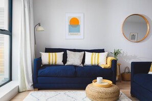 Ein Wohnzimmer mit einem dunkelblauen Sofa, einem runden Spiegel, einem gerahmten Kunstwerk und einem kleinen, runden Tisch mit einer Tasse.