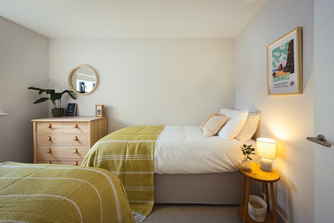 Ein Schlafzimmer mit einem Bett, einem Nachttisch mit Lampe, einer Kommode, einem Spiegel und einem gerahmten Bild an der Wand.