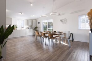 A modern kitchen with white cabinets, a dining table with chairs, and a large window providing natural light. - 4 Bed in Falmouth (oc-b588809) (Goldenbank)