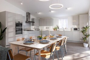 A modern kitchen with built-in appliances, a dining area with a long table set for six, and a bright, airy atmosphere. - 4 Bed in Falmouth (oc-b588809) (Goldenbank)