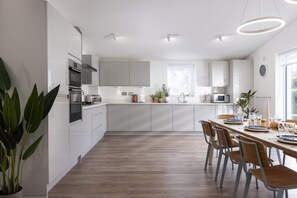 A modern kitchen with white cabinetry, a dining area with wooden chairs, and a large window providing natural light. - 4 Bed in Falmouth (oc-b588809) (Goldenbank)