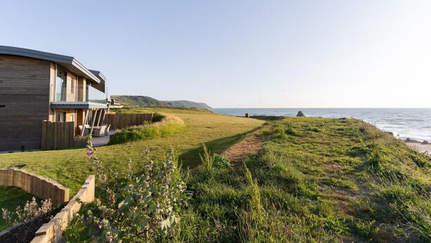 A modern house with a deck overlooking a grassy area and the ocean. - 5 Bed in Widemouth Bay (oc-b628538) (Widemouth Bay)