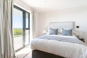 A bedroom with a large bed, a headboard, two bedside lamps, and a view through a sliding glass door. - 5 Bed in Widemouth Bay (oc-b628538) (Widemouth Bay)