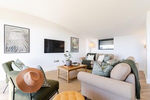 A modern living room with a sofa, armchair, and coffee table, featuring artwork on the walls and a television. - 5 Bed in Widemouth Bay (oc-b628538) (Widemouth Bay)