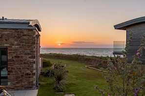 A sunset view from a stone building with a grassy yard and a wooden fence. - 5 Bed in Widemouth Bay (oc-b628538) (Widemouth Bay)