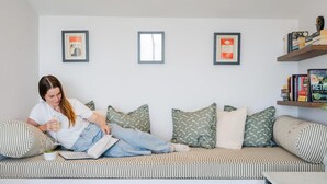 A person sitting on a striped sofa with a book, a cup, and a bowl of fruit. There are pillows, a shelf with books, and framed pictures on the wall. - 5 Bed in Widemouth Bay (oc-b628538) (Widemouth Bay)