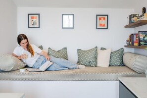 Eine Person sitzt auf einem gestreiften Sofa, ein Buch, eine Tasse und eine Schale mit Obst in der Hand. Kissen, ein Bücherregal und gerahmte Bilder sind an der Wand zu sehen.