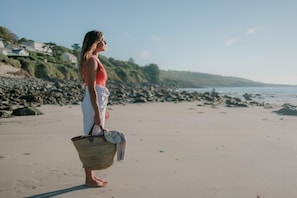 A person standing on a sandy beach with a rocky shoreline and houses in the background. - 4 Bed in The Lizard (oc-b620048) (Coverack)