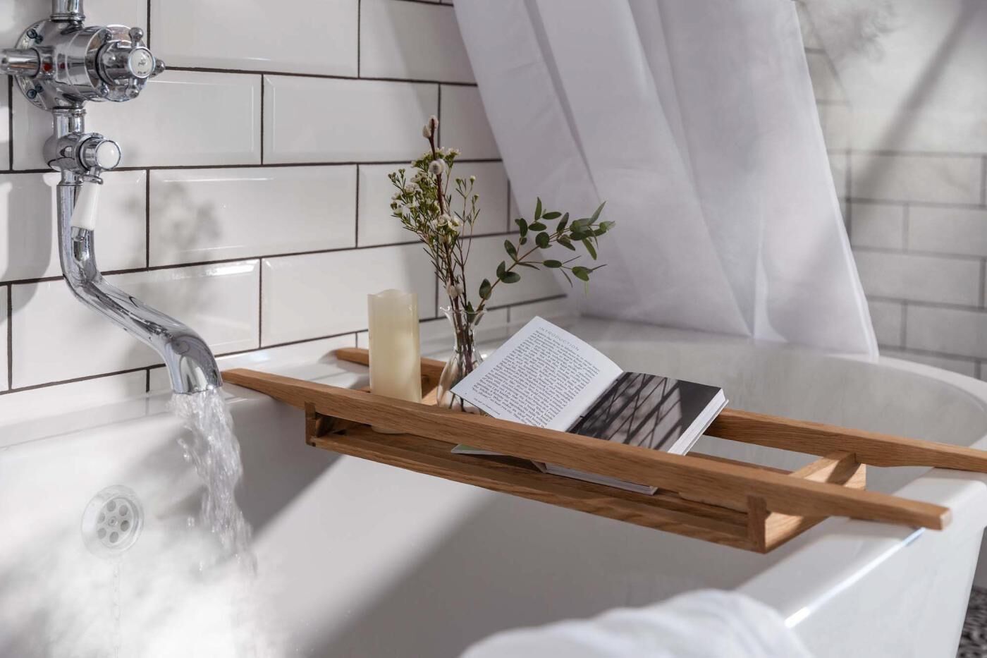 Une salle de bain avec une baignoire, un plateau en bois sur lequel sont posés un livre et une bougie, et un robinet d’où coule de l’eau.