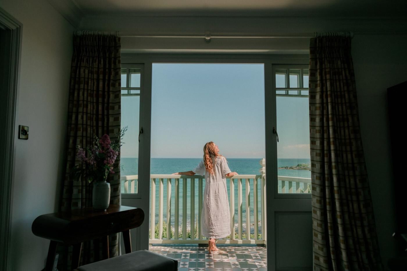 A room with a view of the sea through a balcony door, a chair, and a vase with flowers.
