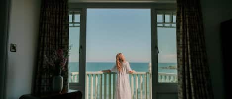 A room with a view of the sea through a balcony door, a chair, and a vase with flowers.