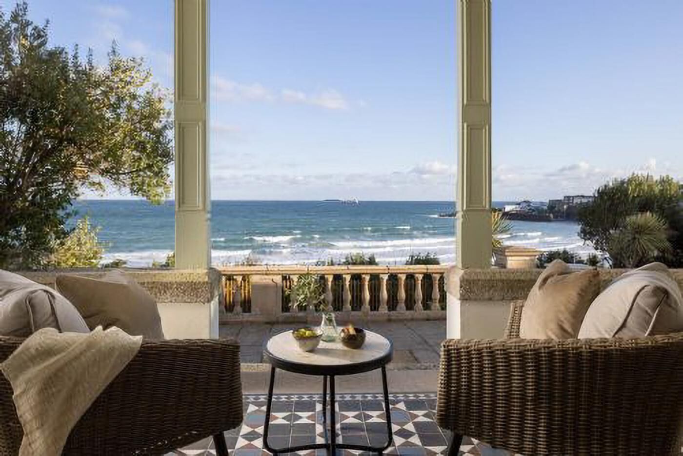 Een balkon met rieten meubilair en een tafeltje, met uitzicht op een strand met golven en verre gebouwen.
