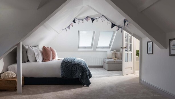 A bedroom with a sloped ceiling, a bed with pillows, a bench, and a view of the outside. - 4 Bed in The Lizard (oc-b620048) (Coverack)