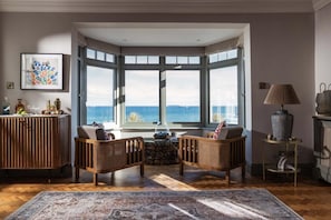 A room with a bay window overlooking the sea, featuring wicker furniture, a rug, and a side table with a lamp. - 4 Bed in The Lizard (oc-b620048) (Coverack)