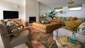 A living room with a sofa, armchair, ottoman, and a flat-screen TV. - 4 Bed in Watergate Bay (oc-b584345) (Tregurrian)