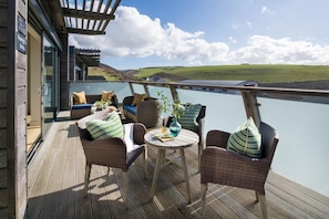 A deck with wicker furniture, a table, and a view of a lake. - 4 Bed in Watergate Bay (oc-b584345) (Tregurrian)