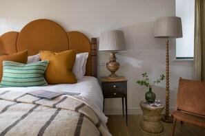 A neatly made bed with a headboard, a bedside table with a lamp, a small side table with a vase, and a chair. - 4 Bed in Watergate Bay (oc-b584345) (Tregurrian)